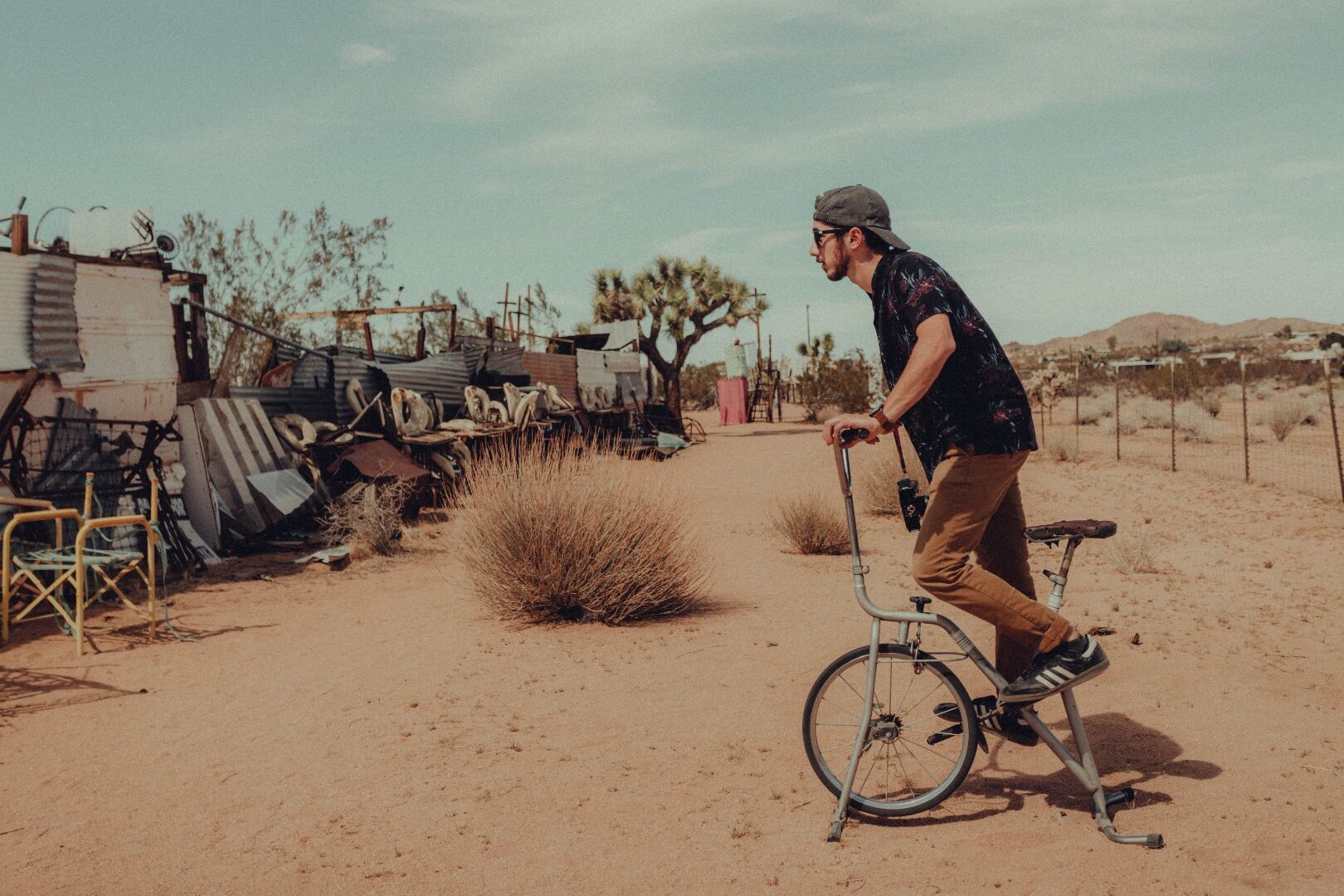 A man riding a stationary bike through a desert lot in Joshua Tree, with weathered metal and brush around him.