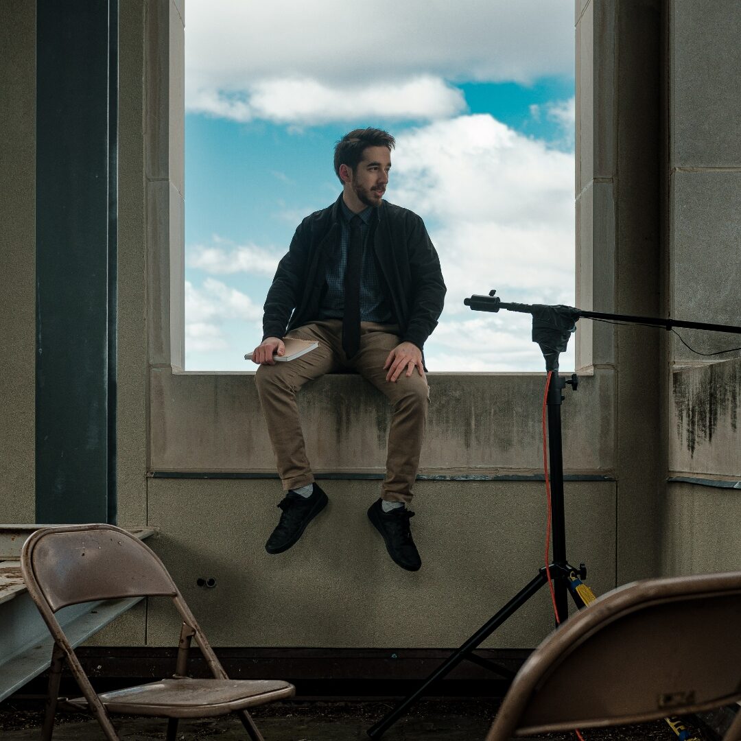 A man seated in a tall open window above empty folding chairs, looking outward toward a bright sky.